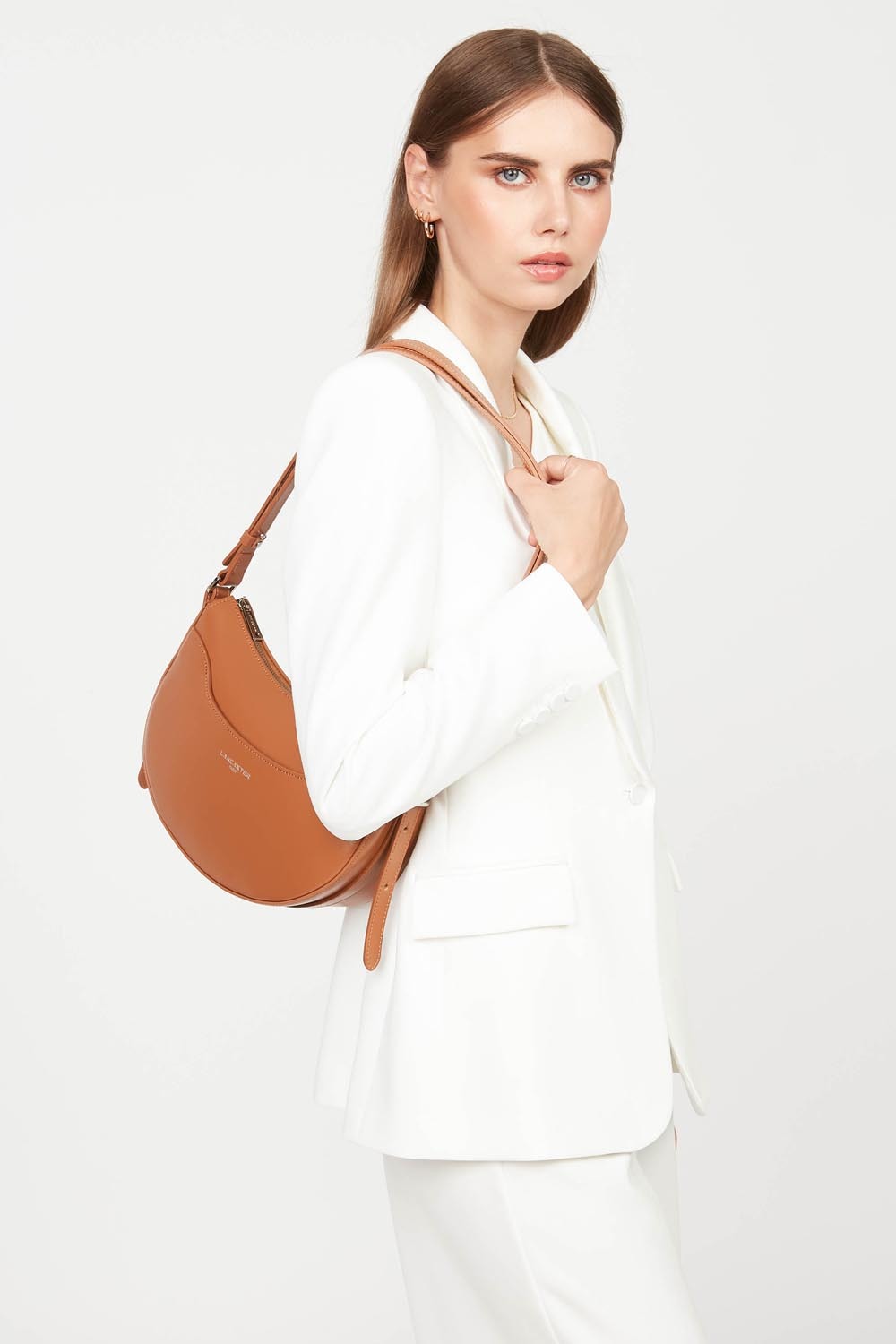 Lancaster besace Suave Ace marron portée en bandoulière avec un tailleur blanc.