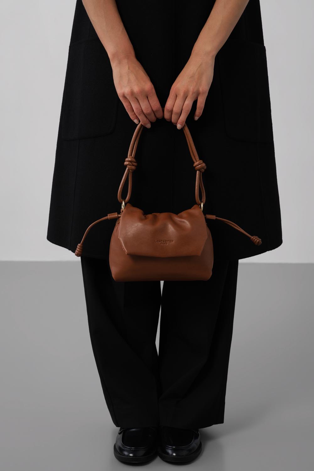 Lancaster besace Lush marron portée à la main devant une tenue noire.