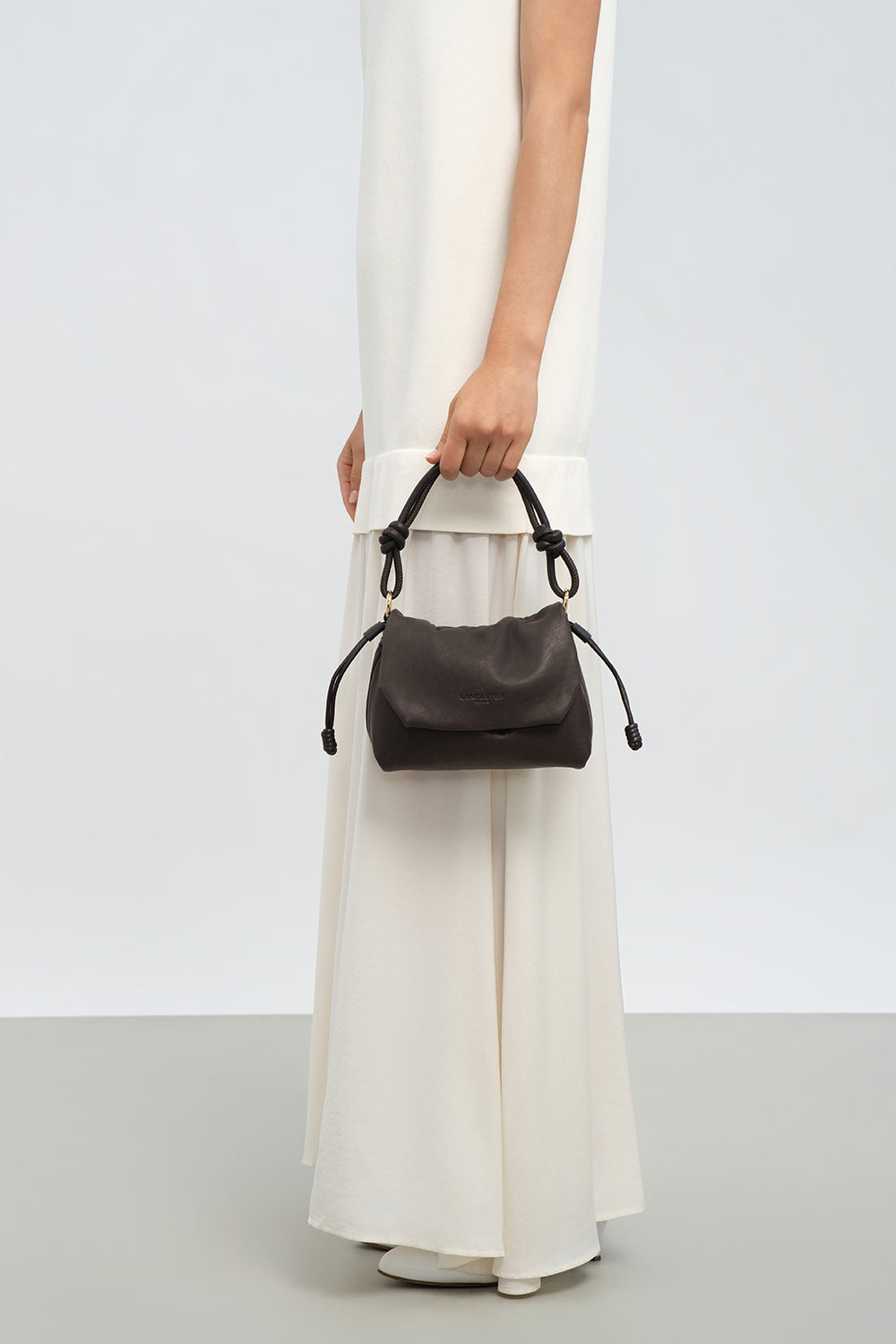 Lancaster besace Lush marron tenue à la main, avec une longue robe blanche.